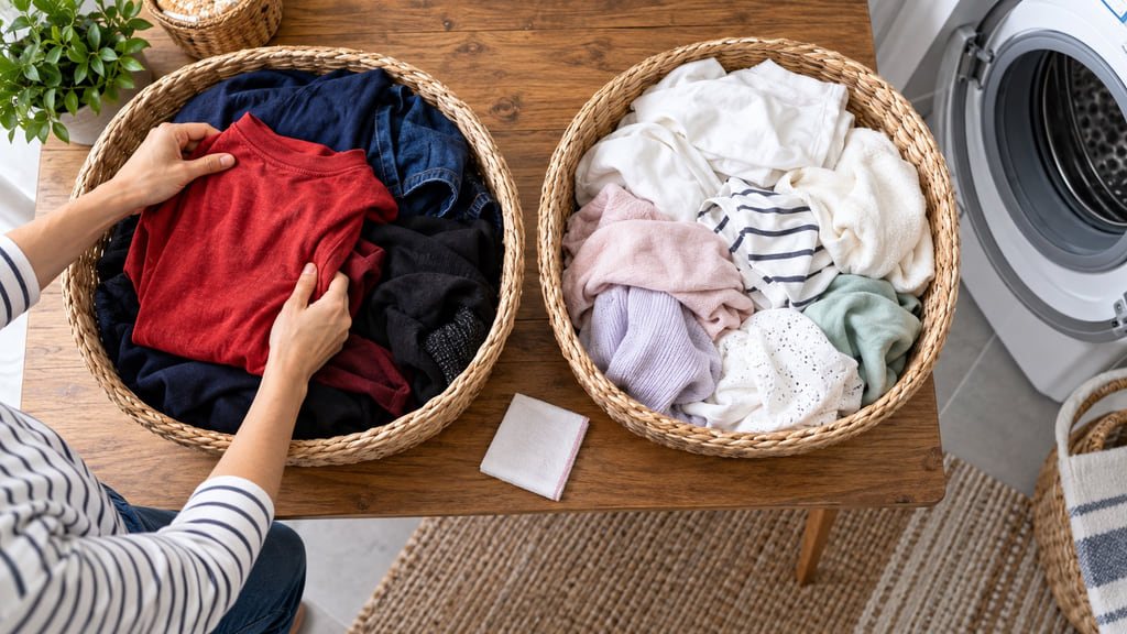 Two laundry baskets separating a red shirt and dark clothes from white and pastel garments beside a washer.