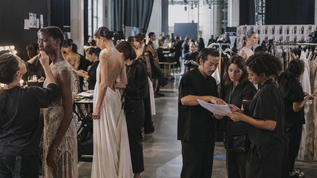 Adult fashion models, stylists, and production staff preparing backstage before a runway show.